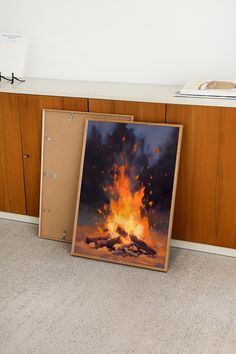 a painting sitting on the floor in front of a cabinet with an open fire burning behind it