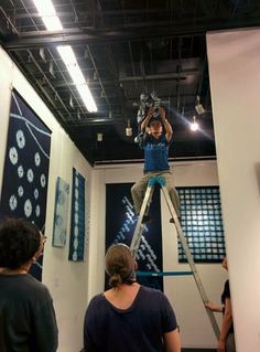 people are standing around and looking at art on display in an exhibit room with ladders