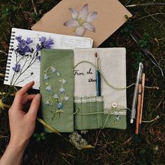 a hand holding a pen next to two notebooks and some flowers on the ground