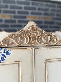 an ornate white cabinet with blue flowers painted on the front and side panels, in front of a brick wall