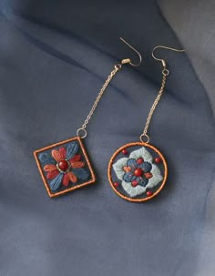 a pair of earrings with flowers on them sitting on a blue cloth surface, one is made from metal and the other is beaded