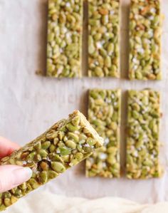 a hand holding a piece of granola bar with nuts and seeds on it, in front of four other bars