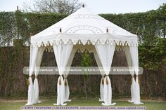 a white gazebo with curtains on it in front of a fence and bushes behind it