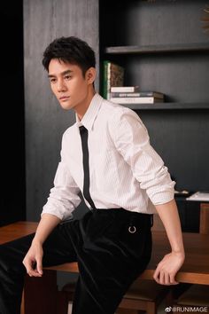 a young man wearing a white shirt and black tie sitting on a wooden table in front of a bookcase