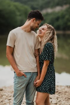 a young man and woman standing next to each other on the shore of a lake