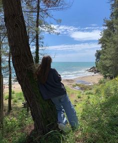 a woman is sitting in the shade of a tree by the beach looking out at the ocean