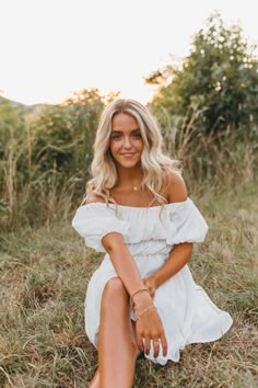 a woman sitting in the grass with her legs crossed, wearing a white dress and sandals