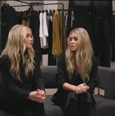 two women sitting on a couch talking to each other in front of some clothes racks