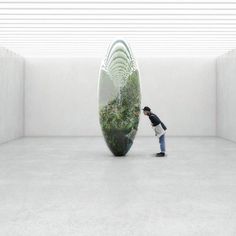 a woman standing in front of a large mirror with plants inside it and another person taking a photo