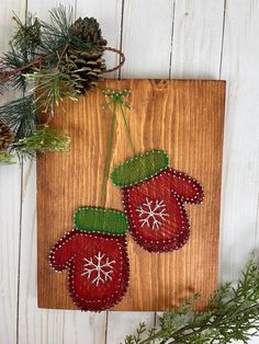 two red mittens are hanging on a wooden board next to pine cones and evergreen branches