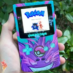 a person holding up a gameboy in front of some bushes and trees, with the screen showing pokemon on it