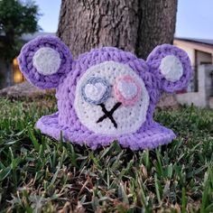 a crocheted teddy bear laying in the grass next to a tree with its eyes closed