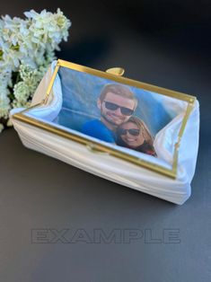 a white and gold purse with a photo on the front is sitting next to some flowers