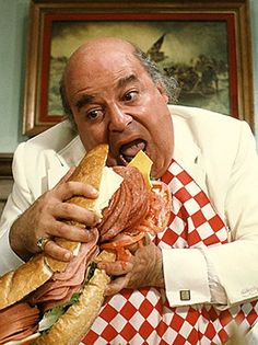 an older man eating a giant sandwich with meat and cheese in his hands while sitting on a chair