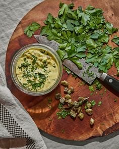 a wooden cutting board topped with greens next to a knife and bowl of soup on top of a table