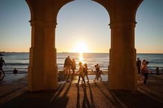 people walking on the beach at sunset under archways with water and sky in background