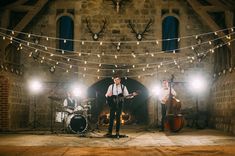 three men are playing instruments in an old brick building with string lights strung across the room