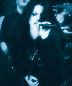 a woman singing into a microphone while standing next to a man with long black hair