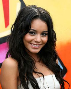 a woman with long black hair and blue eyes smiling in front of a graffiti wall