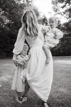 a woman holding two babies in her arms while standing on the grass with trees in the background