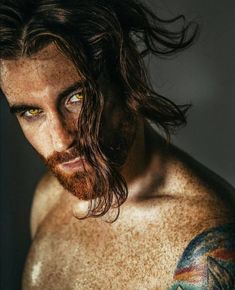 a man with long hair and green eyes is looking at the camera while he has tattoos on his chest