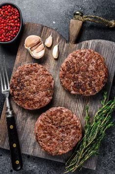 three hamburger patties on a cutting board with garlic and peppercorst next to them