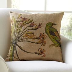 a green bird sitting on top of a pillow next to a white chair in front of a window