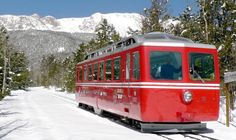 a red trolley car traveling down a snow covered road next to evergreen trees and mountains