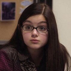 a girl with long brown hair and glasses is looking at the camera while holding a cell phone in her hand