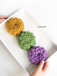 three crocheted flowers sitting in a white box on top of a table next to a person's hand