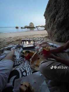 two people are sitting on the beach with food and drinks in front of their laptop