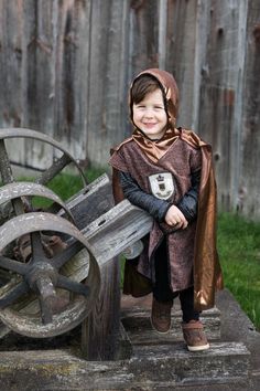 a little boy dressed up in a costume standing next to an old wooden cart with wheels