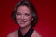 a woman in a black dress smiling at the camera with a red wall behind her