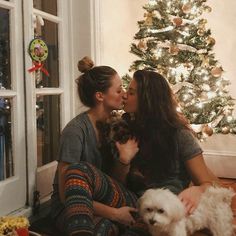 two women kissing while sitting on the floor with their dogs in front of a christmas tree