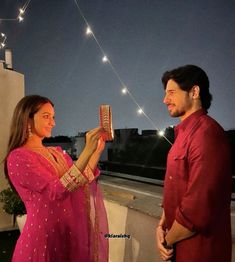 a man and woman standing next to each other on a roof at night with string lights above them