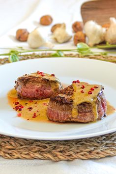 two pieces of steak on a white plate with sauce and nuts in the back ground