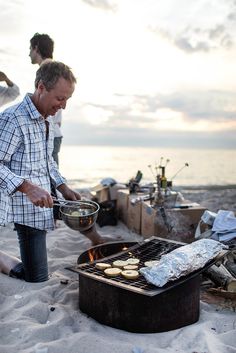 a man grilling food on the beach with his family watching from the water's edge