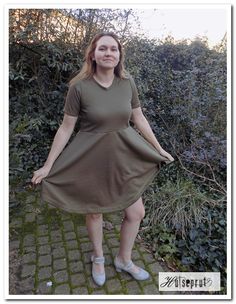 a woman in a green dress standing on a brick walkway next to bushes and shrubbery