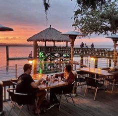 two people sitting at a table on a pier with the sun setting in the background