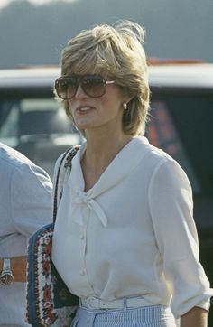 a woman wearing sunglasses and a white blouse is walking in front of a car while holding a purse