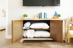 a tv mounted above a wooden cabinet with white pillows and blankets on it's sides