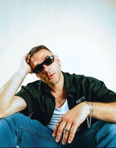 a man wearing sunglasses sitting on the ground with his hands behind his head and looking off to the side