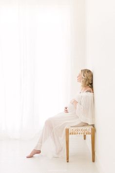 a woman sitting on top of a wooden chair in front of a white wall and window