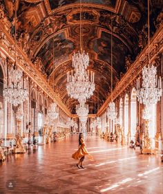 a woman is walking in an ornate hall with chandeliers and paintings on the walls