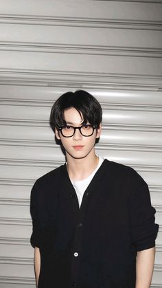 a young man wearing glasses standing in front of a white wall with his hands on his hips