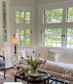 a living room filled with furniture and flowers on top of a coffee table in front of two windows