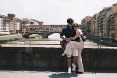 a man and woman are sitting on a wall in front of a river with buildings