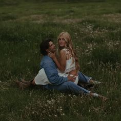 a man and woman sitting on the ground in a field with wildflowers looking at each other