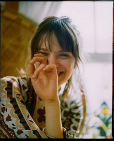 a woman is smiling and holding her hand up to her face while sitting in front of a window