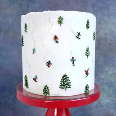 a white frosted cake with trees and skiers on the top is sitting on a red stand